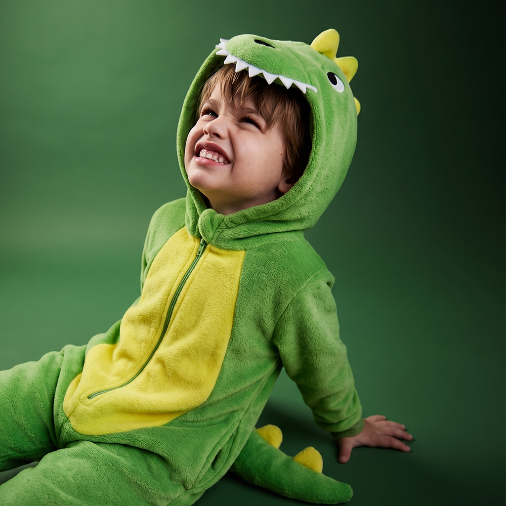 A smiling young child wearing DinoBOO, a plush green dinosaur onesie with a yellow belly panel, yellow horns, and a toothy hood, posed against a dark green studio background.