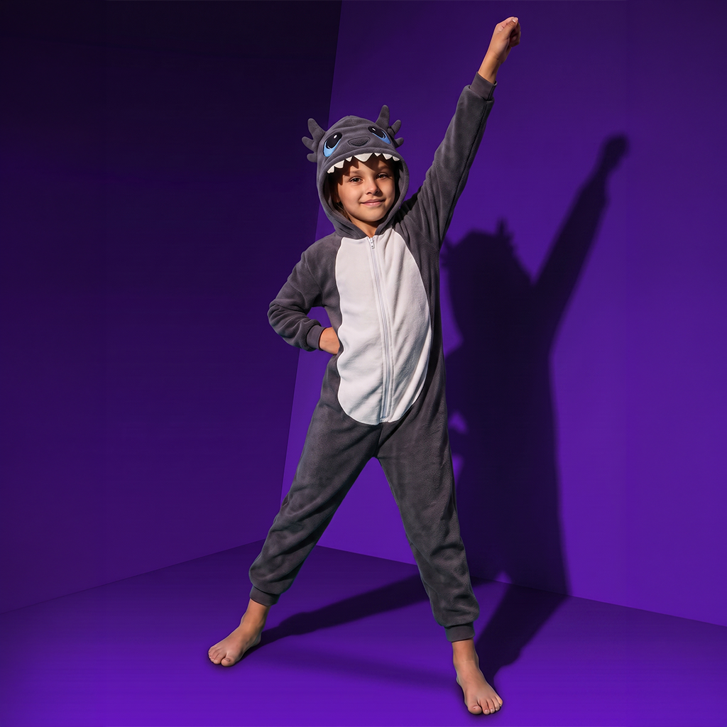 A child modeling GriBOO the grey dragon and white Stitch-themed animal onesie with a hood, standing against a purple studio background with a strong shadow cast on the wall.