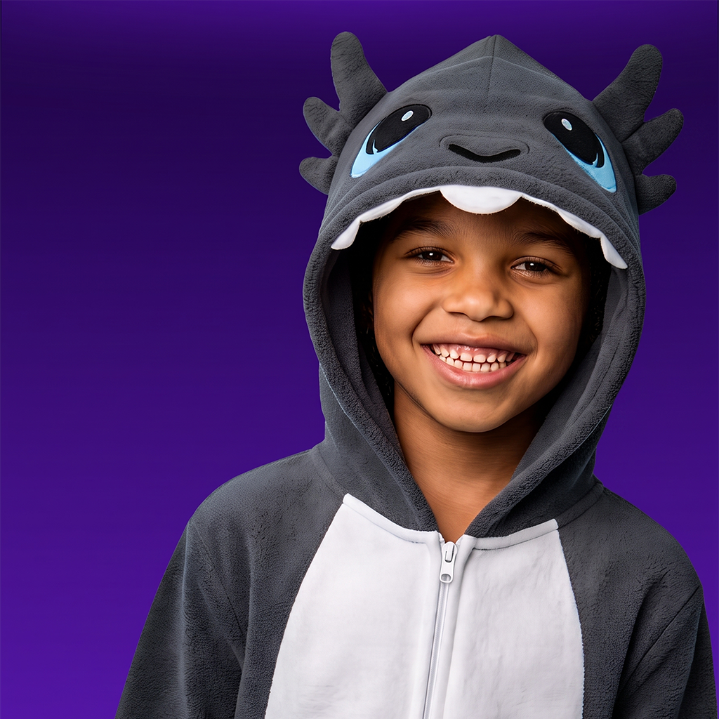 A smiling child wearing GriBOO the grey dragon a plush hooded zip-up costume with a dark grey and white body, blue eyes on the hood, and horn-like protrusions, photographed against a solid purple studio background.